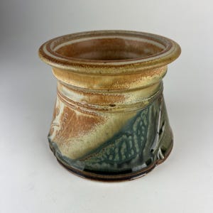 May include: A handcrafted ceramic pot with a wide, rounded rim. The pot features a blend of earthy tones, including brown, green, and beige. The exterior has textured details and a glossy finish, suggesting a unique, artisanal design. The pot is suitable for decorative or functional use.