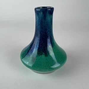 Puede incluir: Un jarrón pequeño y bulboso con cuello estrecho, que presenta un esmalte liso y brillante que transiciona desde un azul marino profundo y oscuro en la parte superior hasta un verde esmeralda vibrante y brillante en la base.