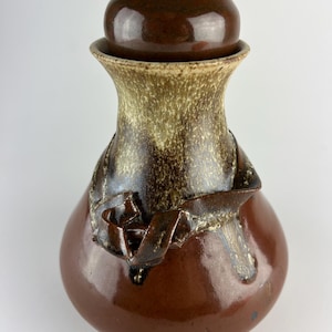 May include: A handcrafted ceramic jar with a lid. The jar is a rich brown color with a lighter tan and brown band around the neck. A decorative brown ribbon-like design wraps around the body. The lid is a rounded shape.