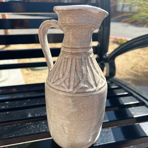 May include: A beige ceramic pitcher with a handle and spout. The pitcher has a carved, textured design around the neck and body. The pitcher is placed on a black bench. The pitcher is approximately 20 cm tall.