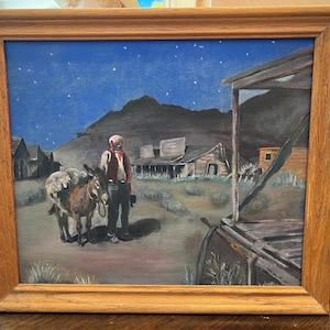 May include: A framed oil painting depicting a desert scene at night with a person standing next to a donkey carrying a pack. The landscape features rustic wooden buildings, sparse vegetation, and a dark blue sky filled with small white stars.