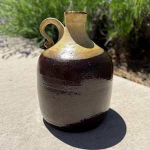 May include: A stoneware jug featuring a glossy dark brown glaze on the lower body and a contrasting light beige glaze on the upper neck and shoulder. The vessel has a single curved handle attached to the side and a narrow, circular opening.