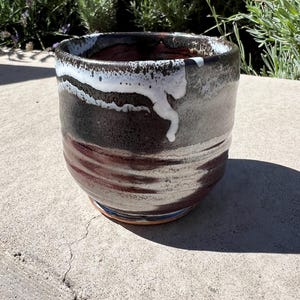 以下が含まれることがあります： 丸みを帯びた形状の陶器製カップ。ダークブラウンとブラックのベース釉薬に、縁から垂れ落ちるような厚みのある白いドリップ模様が施されています。表面は質感があり、胴体部分には水平方向の筋がはっきりと見えます。