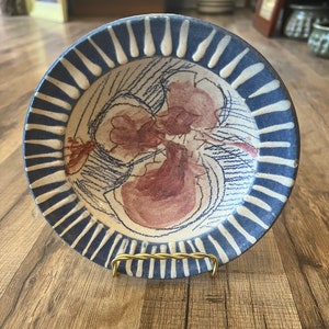 May include: A small ceramic plate with a blue and white striped border and a hand-painted design of a flower in shades of pink and red.