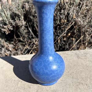 May include: A blue ceramic vase with a rounded base and a slender neck. The vase has a textured surface and a slightly flared rim. The vase is on a concrete surface.