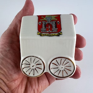 May include: A white ceramic carriage-shaped dish with gold trim and wheels. The top features a crest with the text "NEVER SHALL WONNE-NE-NEVER REDHILL". The dish is held in a hand.