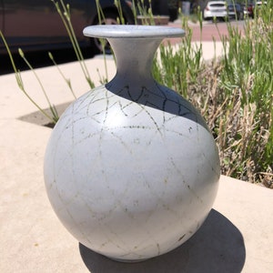 May include: A light gray ceramic vase with a round body and a narrow neck. The vase has a delicate, hand-painted design of thin lines and dots.