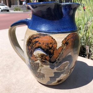May include: A ceramic pitcher with a blue glaze and a brown, black and white abstract design. The pitcher has a wide mouth and a handle.