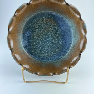 Può includere: Un piatto in ceramica con bordo smerlato, caratterizzato da una sfumatura di colori. Il centro è blu scuro, che sfuma in un verde acqua macchiettato, e poi in un bordo marrone. Il piatto è esposto su un supporto metallico dorato.