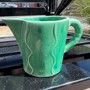 Puede incluir: Una jarra de cerámica verde con un diseño ondulado. La jarra tiene un asa y una boca ancha.