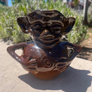 May include: A brown ceramic figurine of a goblin with a closed-eye expression. The goblin is holding a round object in its hands.