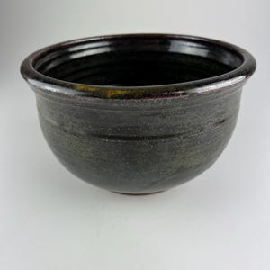 May include: A handcrafted ceramic bowl with a dark, mottled glaze. The bowl has a rounded shape with a slightly flared rim. The colours include shades of black, green, and brown. The bowl is likely used for serving or display.
