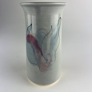 Op de afbeelding: Cilindervormige keramische vaas in lichtblauw met een handgeschilderd ontwerp. De vaas heeft abstracte vormen in blauw, rood en zwart. Het heeft een cilindrische vorm met een uitlopende rand. Een decoratief item voor in huis.