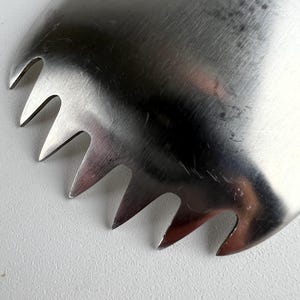 以下が含まれることがあります： 一連の尖った歯が付いたステンレス製のキッチン用品のクローズアップ。この器具は、湾曲した楕円形をしており、つや消し金属仕上げです。歯は等間隔に配置されており、特定の料理の目的に合わせて設計されています。