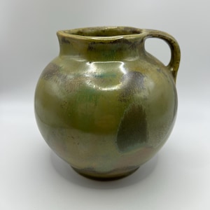 May include: A round ceramic pitcher with a glossy finish, featuring a mottled blend of olive green, earthy brown, and muted tan tones. The vessel has a short, wide neck, a curved handle on the right side, and a smooth, rounded body shape.