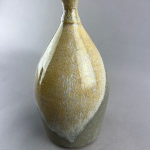 Può includere: Un piccolo vaso in ceramica con una smaltatura marrone chiaro e grigia. Il vaso ha un corpo arrotondato e un collo stretto.