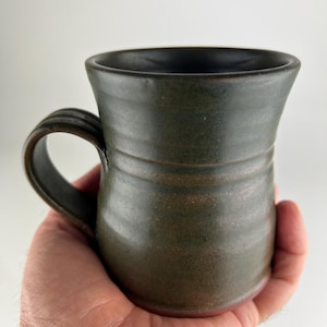 May include: A dark olive green ceramic mug with a flared rim and a rounded body featuring horizontal ridges. The mug has a thick, curved handle on the side and a smooth, dark interior. The base of the mug shows a hint of reddish-brown clay.