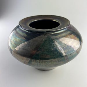 Può includere: Un vaso in ceramica fatto a mano con una finitura scura e iridescente. Il vaso ha un corpo arrotondato e un'ampia apertura circolare. Motivi geometrici in argento e verde adornano la superficie, aggiungendo interesse visivo a questo pezzo decorativo.