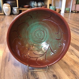 May include: A large, round, ceramic bowl with a green glaze and brown rim. The bowl has a swirling design in the center and a decorative pattern around the rim.