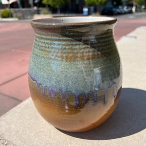 May include: A handmade ceramic mug with a unique glaze. The mug features a rounded body with a wide opening and a textured surface. The colors transition from a warm brown at the base to blue and green, with purple drips. The rim is a darker brown.