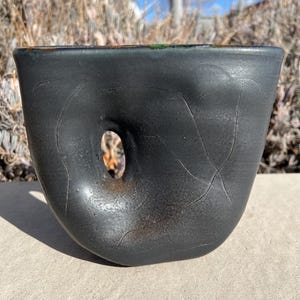 May include: A black ceramic vase with a unique, abstract shape and a hole in the side. The vase has a textured surface and a slightly green rim.