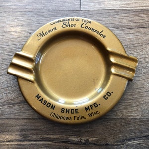May include: A gold metal ashtray with the text "COMPLIMENTS OF YOUR Mason Shoe Counselor MASON SHOE MFG. CO. Chippewa Falls, Wisc."