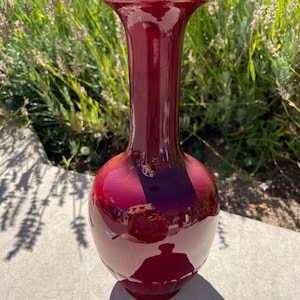 Puede incluir: Un jarrón de cerámica rojo oscuro con cuerpo redondeado y cuello alto y delgado. El jarrón tiene un acabado brillante y una abertura ligeramente acampanada.