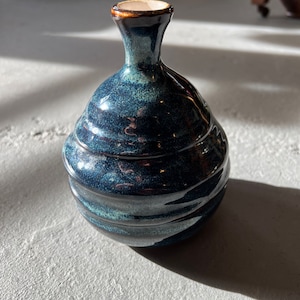 Pode incluir: Um pequeno vaso de cerâmica azul escuro com corpo arredondado e pescoço estreito. O vaso tem um esmalte salpicado e um acabamento ligeiramente iridescente.