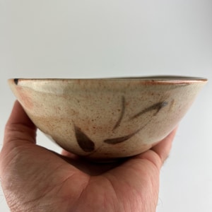 Puede incluir: Un cuenco de cerámica hecho a mano con un exterior beige moteado y diseños marrones en forma de hoja. El cuenco tiene una forma ligeramente irregular y un borde marrón rojizo. El cuenco se sostiene en una mano.