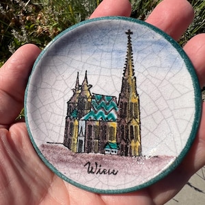Puede incluir: Un plato pequeño y redondo de cerámica con una ilustración pintada a mano de una catedral. El plato tiene un fondo blanco con efecto craquelado, un borde verde turquesa y la palabra "Wien" escrita en negro. La catedral tiene una aguja alta y un tejado verde.