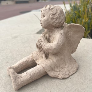 Puede incluir: Una escultura de arcilla beige de un ángel con alas sentado con las piernas cruzadas. El ángel tiene una dulce expresión en su rostro y tiene las manos juntas frente a su pecho.