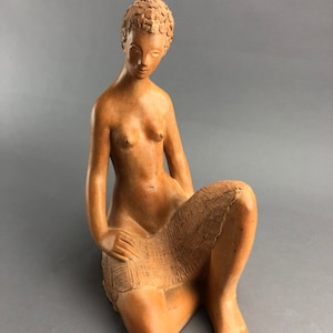 May include: A terracotta-colored sculpture of a seated figure. The figure is nude, with short, textured hair and a serene expression. The sculpture's pose is relaxed, with one leg bent and the other extended.
