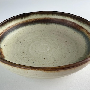 May include: A ceramic bowl with a wide, shallow design. The interior is a speckled cream color, with a dark brown band around the rim and a lighter brown band inside. The rim is a darker brown.