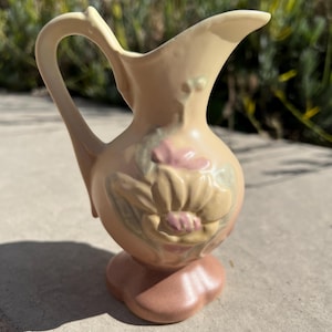 Può includere: Un piccolo brocca in ceramica vintage nei toni del pesca, beige e rosa. La brocca presenta un motivo floreale sul davanti e un manico curvo. La base è di una tonalità di rosa più scura. La brocca è alta circa 13 cm.