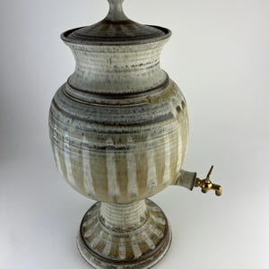 Puede incluir: Un dispensador de agua de cerámica con tapa y grifo de latón. El dispensador es de color blanquecino con rayas verticales y una base de pedestal. La tapa y la base tienen un borde marrón oscuro. El dispensador es probablemente hecho a mano.