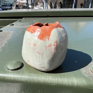 May include: A ceramic vase with a white glaze and a brown rim. The vase has an irregular shape and a textured surface.