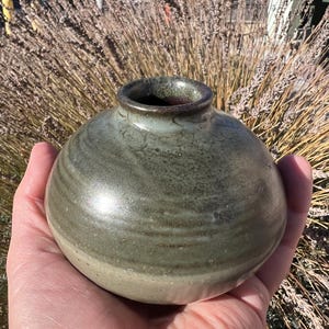 Könnte beinhalten: Kleine, runde Keramikvase mit breiter Öffnung. Die Vase hat eine gedämpfte Grün- und Beigetönung mit sichtbarer Textur und glänzendem Finish. Das handgefertigte Töpferstück wird in einer Hand gehalten, mit einem verschwommenen Hintergrund aus getrockneten Pflanzen.