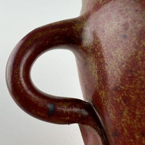 May include: Close-up of a handmade ceramic mug with a large, curved handle. The mug has a speckled, reddish-brown glaze with hints of yellow and black. The handle is a darker shade of reddish-brown.