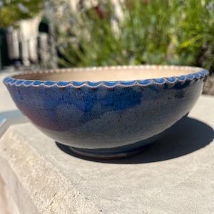 Puede incluir: Un cuenco de cerámica hecho a mano con un borde festoneado. El cuenco tiene un exterior azul oscuro con una textura moteada y un interior beige. El cuenco está colocado sobre una superficie de hormigón, con vegetación en el fondo.