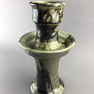 Può includere: Un portacandele in ceramica fatto a mano con un design unico. Il pezzo presenta una struttura a più livelli con una base ampia, una ciotola centrale e una sezione superiore. La tavolozza dei colori comprende sfumature di marrone, nero e crema, con una finitura strutturata.