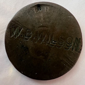 Könnte beinhalten: Nahaufnahme eines dunkelbraunen Metalltokens mit der Gravur "W.B. WILSON" auf der Oberfläche.