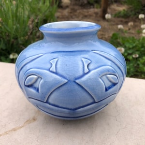 Puede incluir: Un pequeño jarrón de cerámica azul claro con un diseño floral texturizado. El jarrón tiene un cuerpo redondo y una abertura ligeramente acampanada.