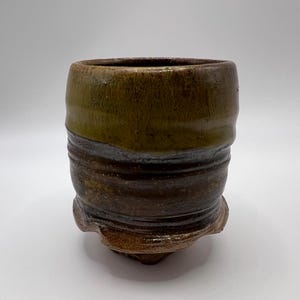 May include: A handcrafted ceramic cup with a distinctive, textured design. The cup has a dark brown base, which fades into a lighter brown and olive green at the top. It has a rounded body and a slightly flared rim, with a textured, organic foot.