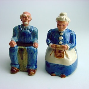 May include: A pair of ceramic figurines depicting a man and a woman. The man is wearing blue overalls and a white shirt. The woman is wearing a blue dress with a white apron and is holding knitting needles. Both figurines have a planter on their laps.