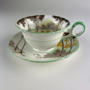 May include: A vintage teacup and saucer set. The white porcelain cup and saucer feature a landscape design with a green trim. The cup's interior displays a forest scene with brown trees and yellow accents. The saucer has a matching design.