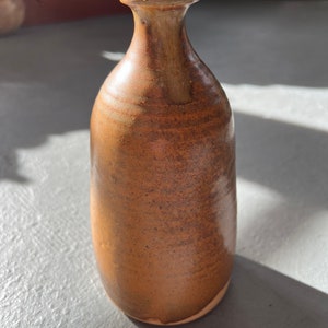 May include: A small, brown ceramic vase with a rounded body and a narrow neck. The vase has a speckled, textured surface.