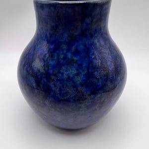 May include: A handcrafted ceramic vase with a rounded body and a flared opening. The vase is glazed in a deep blue color with mottled variations, creating a textured appearance. The overall shape is reminiscent of a classic amphora.