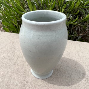 Puede incluir: Un jarrón de cerámica blanco con un esmalte moteado. El jarrón tiene un cuerpo redondeado y un labio acampanado. Está sentado sobre una superficie de concreto.