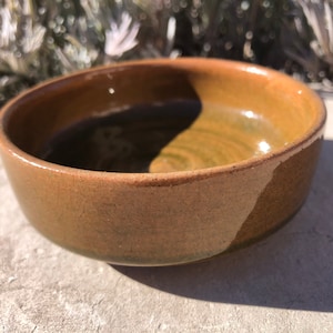 Puede incluir: Un pequeño cuenco redondo de cerámica marrón con un acabado esmaltado. El cuenco tiene un ligero borde alrededor del borde y un sutil patrón de remolino en el interior.