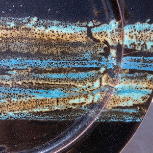 Pode incluir: Um prato de cerâmica preto com um esmalte azul e castanho. O esmalte tem uma textura salpicada e cria um padrão único.